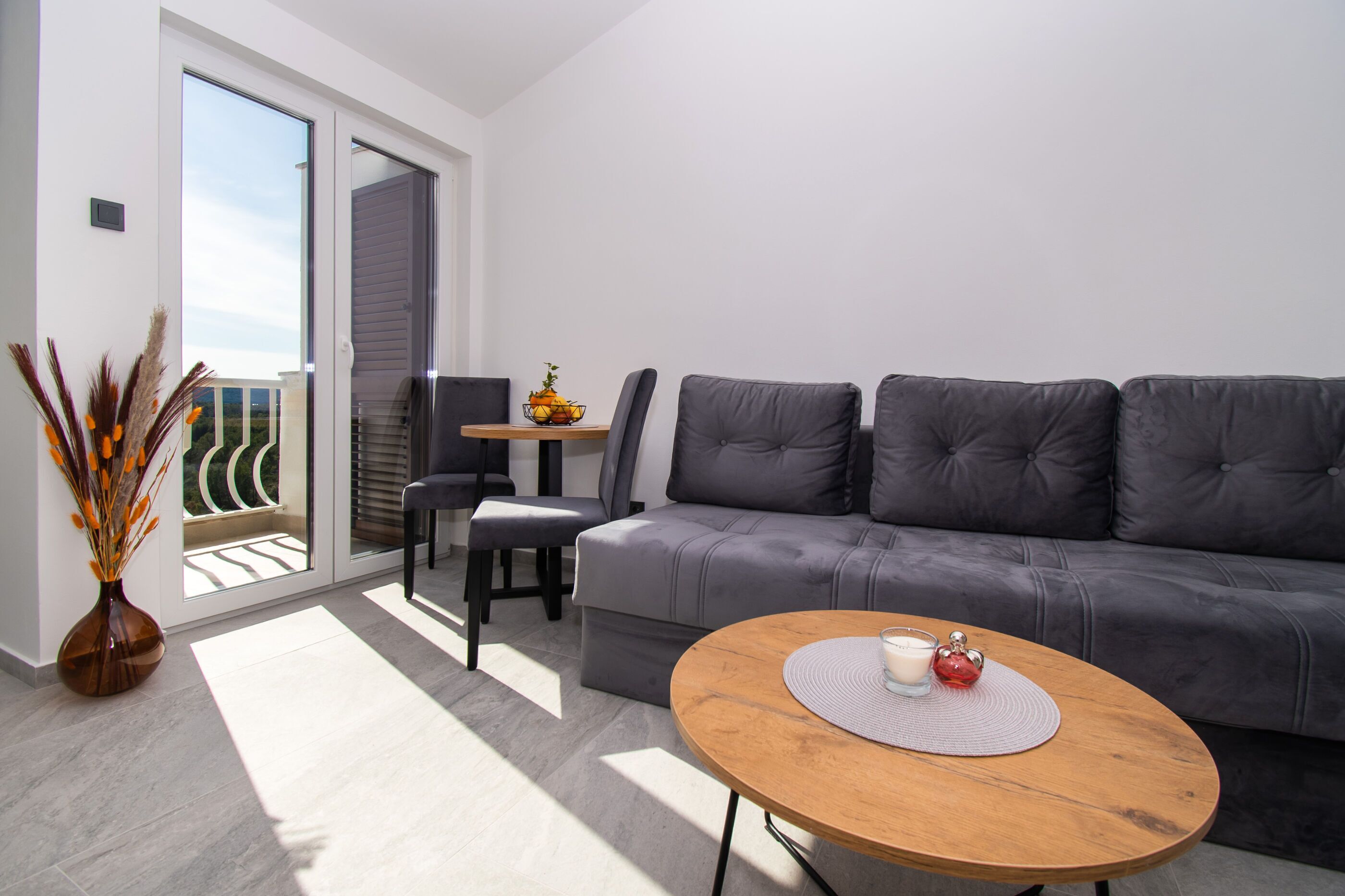 A modern living room with a grey sofa, a wooden coffee table, a dining table with chairs, and a vase with dried plants.