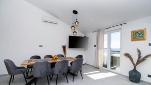 A modern dining area with a wooden table, grey upholstered chairs, a flat-screen TV mounted on the wall, and a large window with a view. - Apartments Valeva Exclusive (Sibenik)