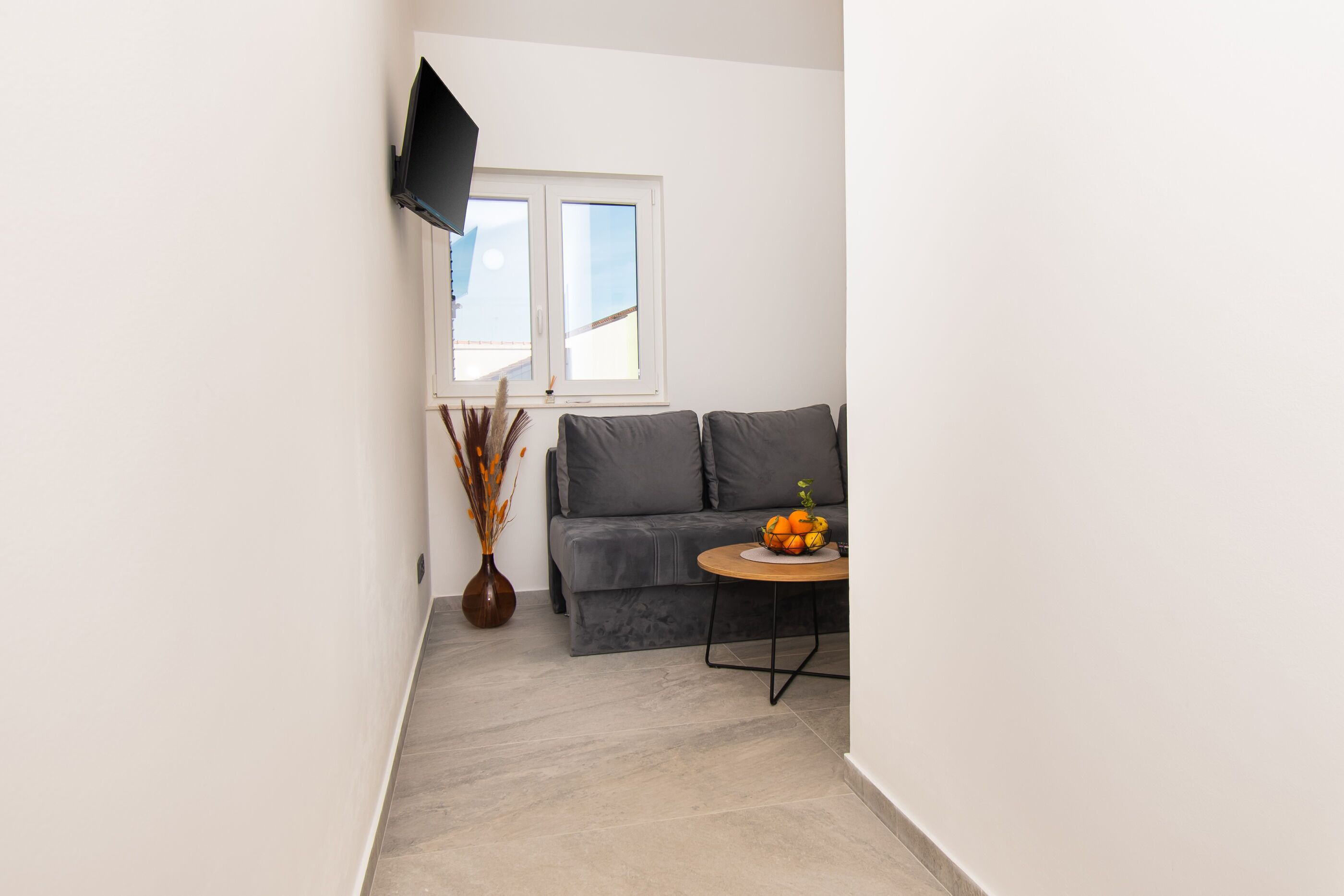 Ein modernes Wohnzimmer mit einem grauen Sofa, einem runden Couchtisch mit Obst, einer Vase mit Trockenpflanzen und einem an der Wand befestigten Fernseher.