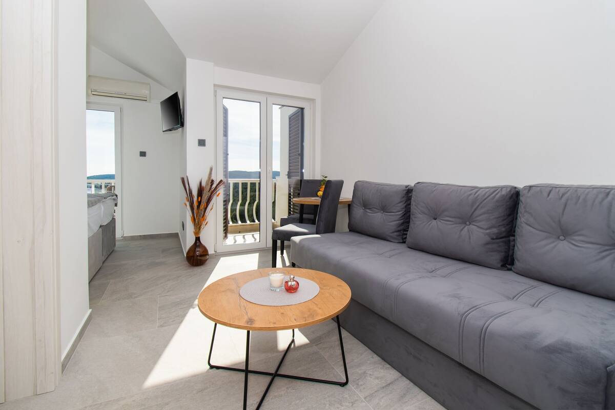 A modern living room with a grey sofa, a wooden coffee table with a candle, and a view of a balcony with a chair.