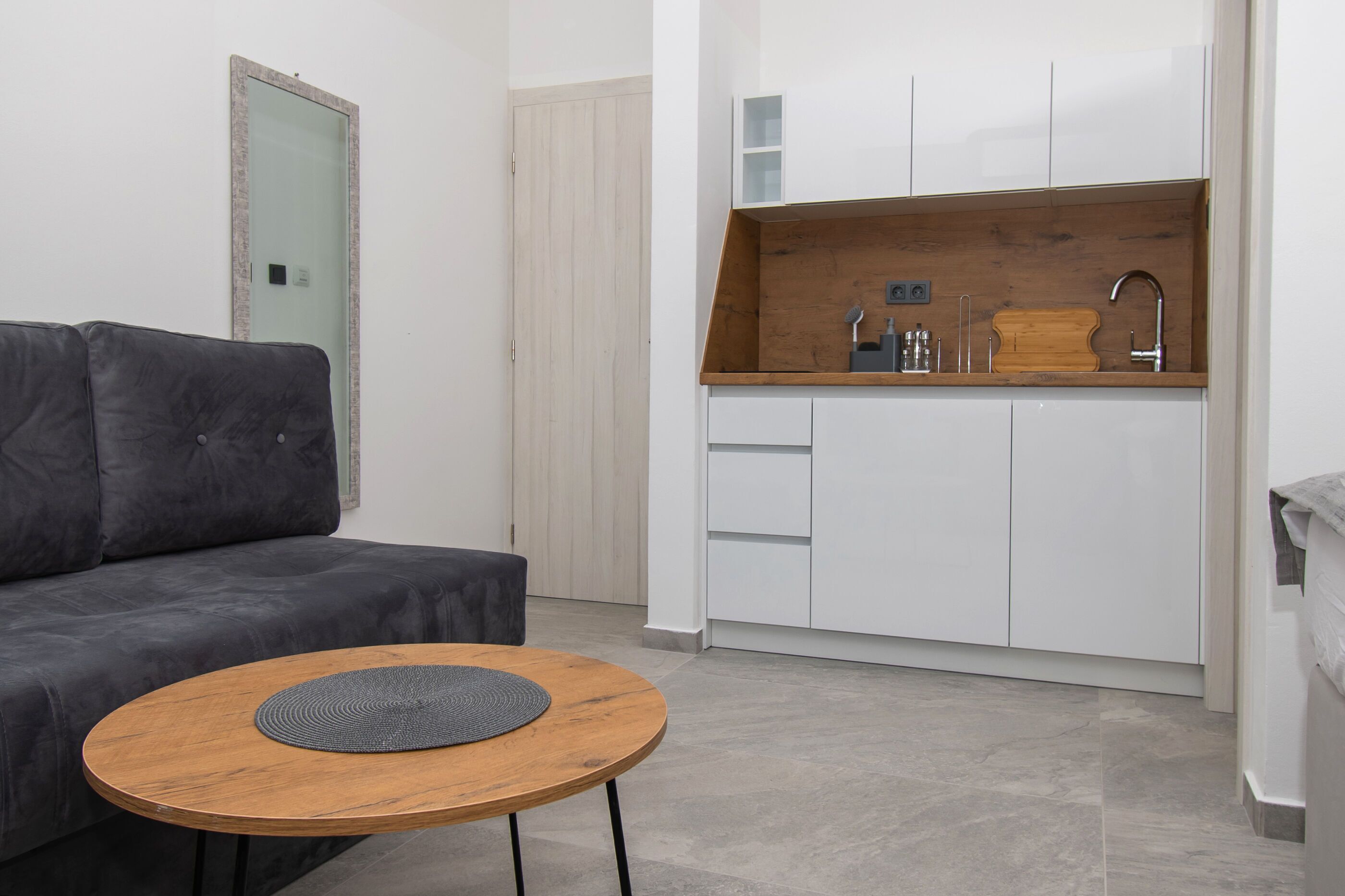 Een knus woongedeelte met een bank, een klein rond tafeltje, een kitchenette en een spiegel.