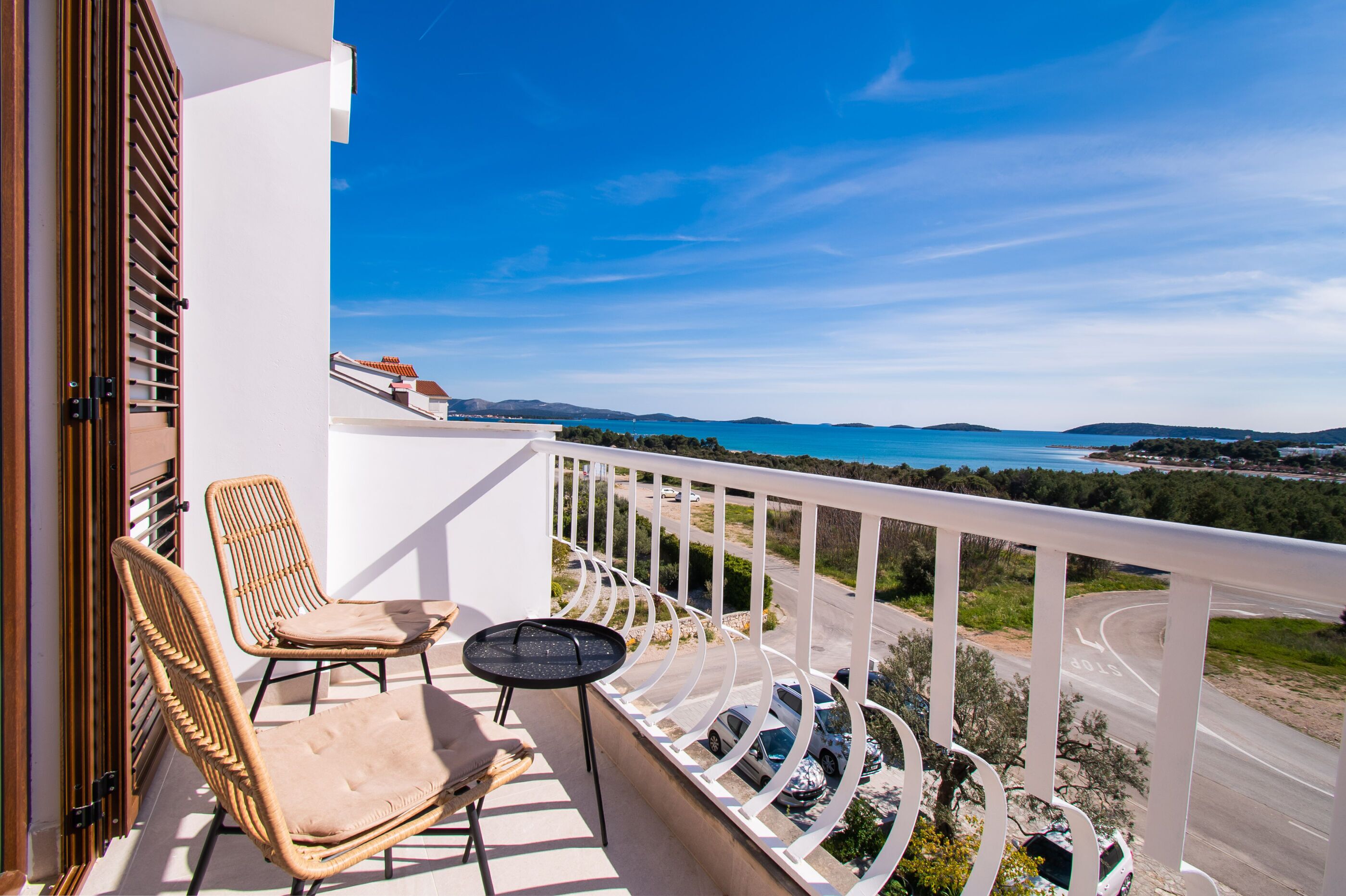 A balcony with wicker chairs and a small round table, overlooking a coastal road with a clear blue sky.