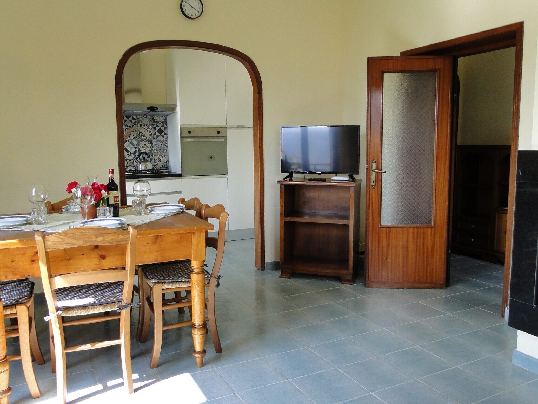 Un coin repas avec une table et des chaises en bois, un téléviseur posé sur un meuble, et une cuisine en arrière-plan.