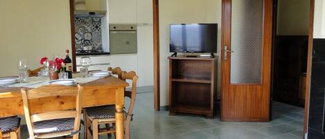 Un coin repas avec une table et des chaises en bois, un téléviseur posé sur un meuble, et une cuisine en arrière-plan.