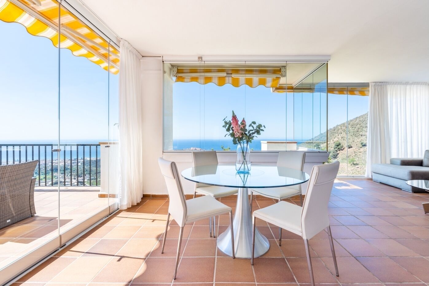 A modern living room with a glass table, white chairs, a sofa, and a view of the sea.