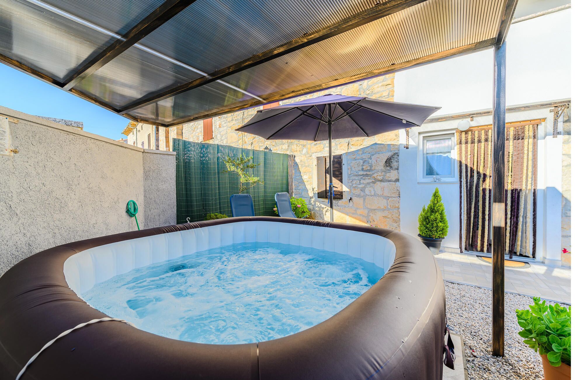 Een hottub onder een overdekt terras, met uitzicht op een stenen gebouw en een groene hekken.