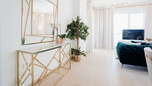 Una sala de estar moderna con una mesa consola de vidrio, un espejo grande, una planta en maceta, un sofá verde oscuro y un televisor de pantalla plana.
