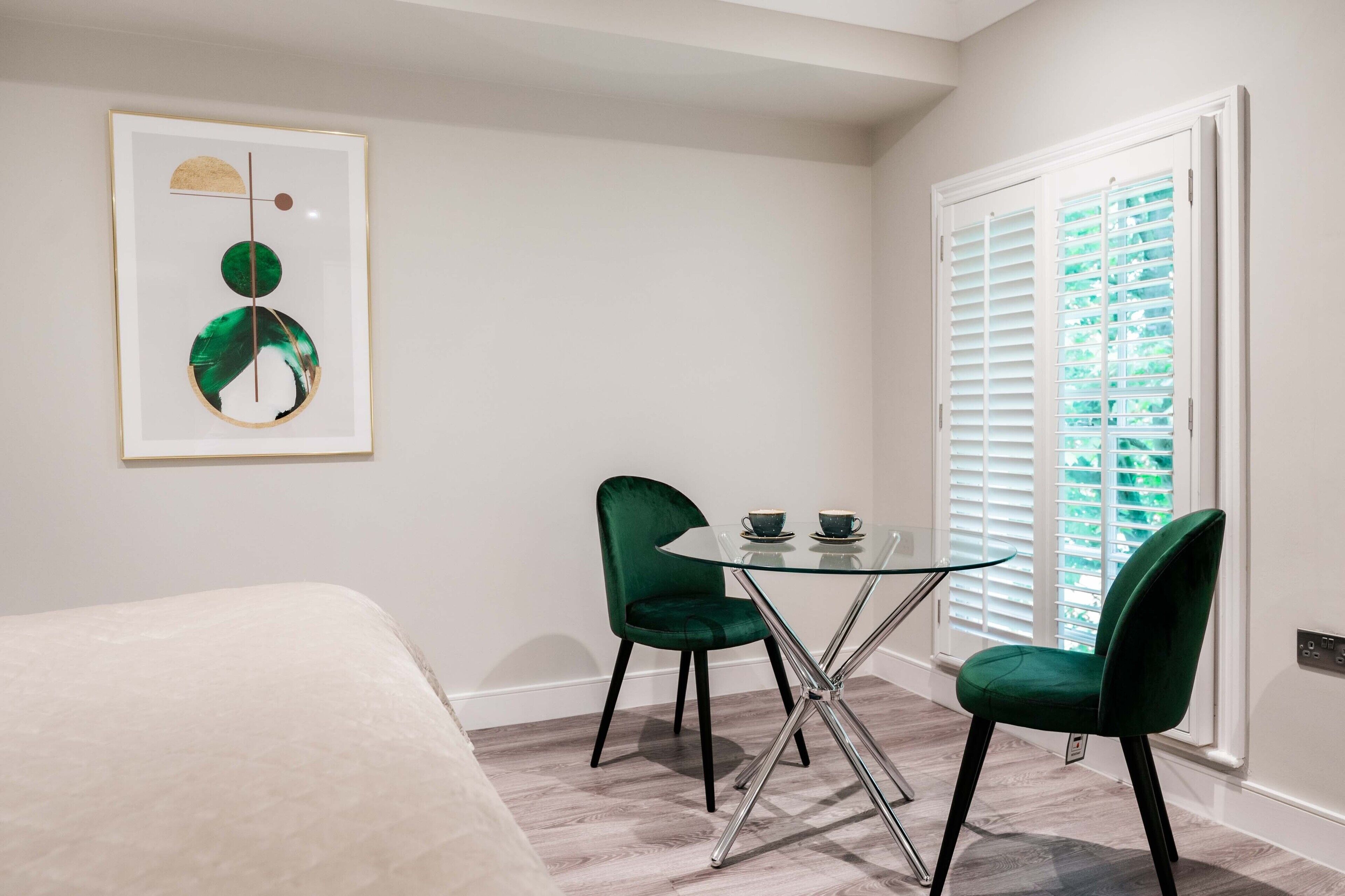 A hotel room with a bed, a glass table with two chairs, a framed artwork, and a window with blinds.