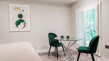 A hotel room with a bed, a glass table with two chairs, a framed artwork, and a window with blinds.