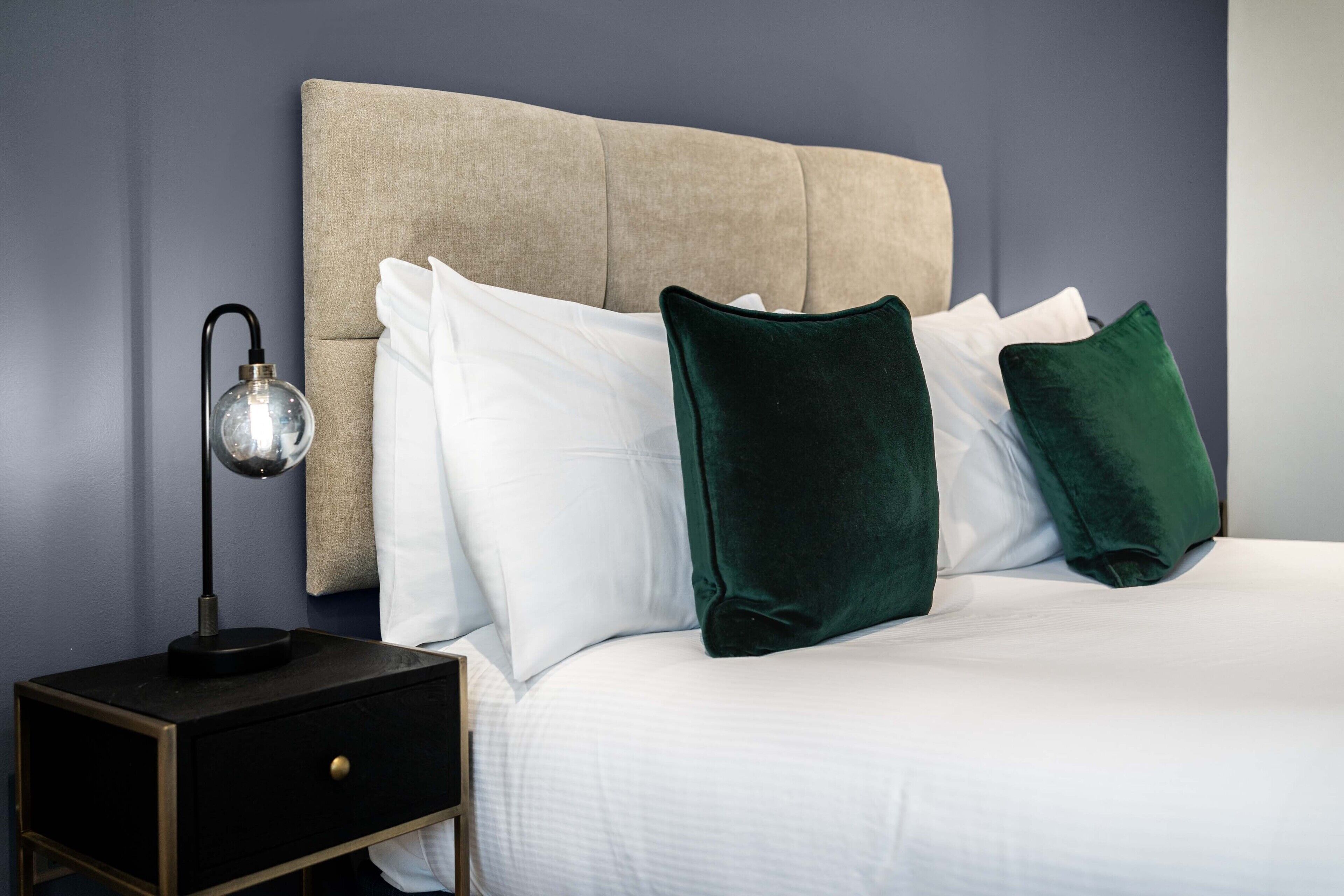 Un letto rifatto con cuscini bianchi e verdi, una testiera beige imbottita e un comodino con una lampada.