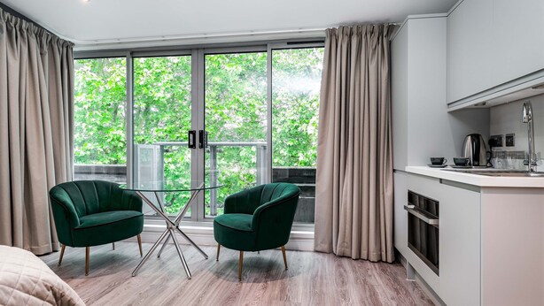 A modern room with a large window, a dining table, two green armchairs, and a kitchen area. - The Regent Aparthotel (Cambridge)