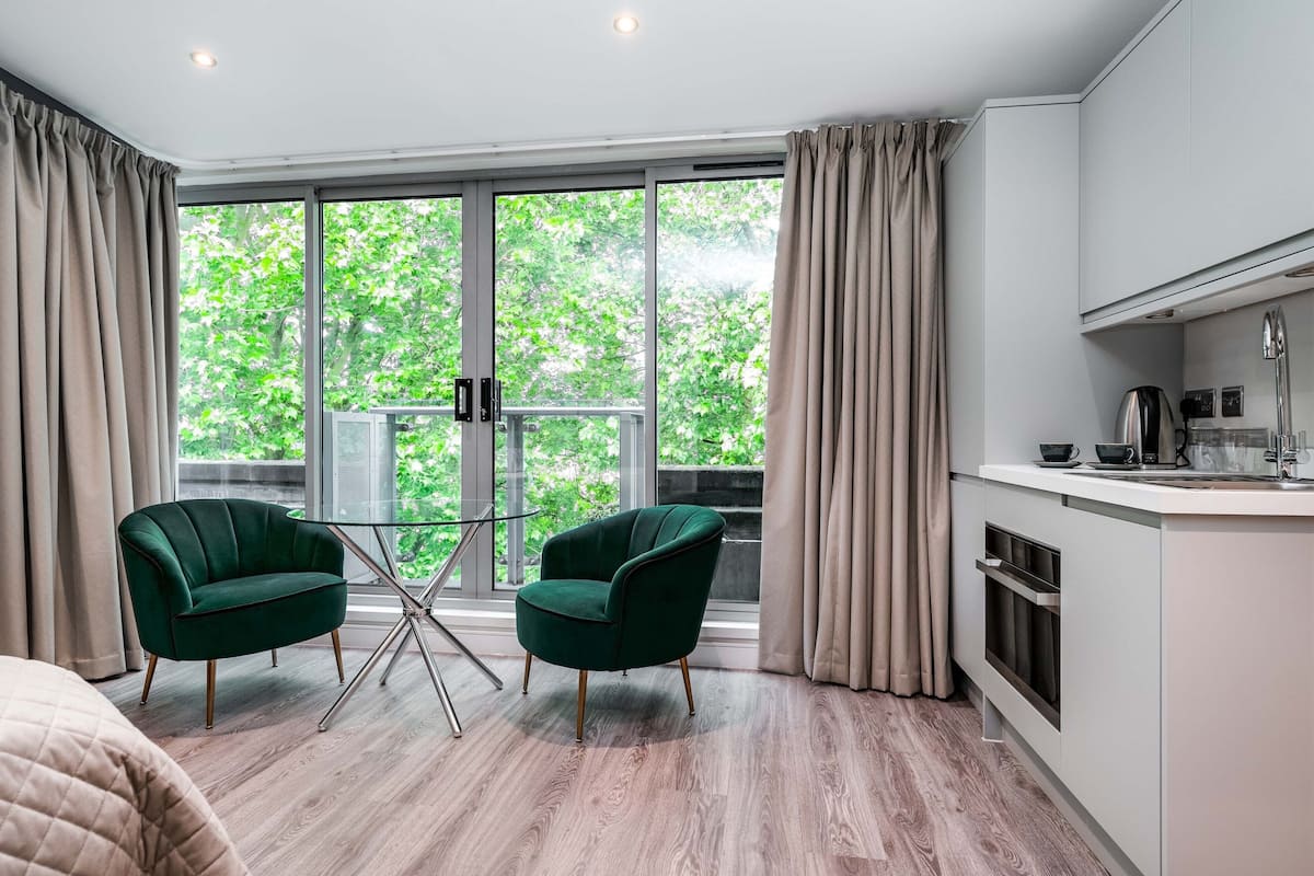 Une pièce moderne dotée d’une grande fenêtre, d’une table à manger, de deux fauteuils verts et d’un coin cuisine.