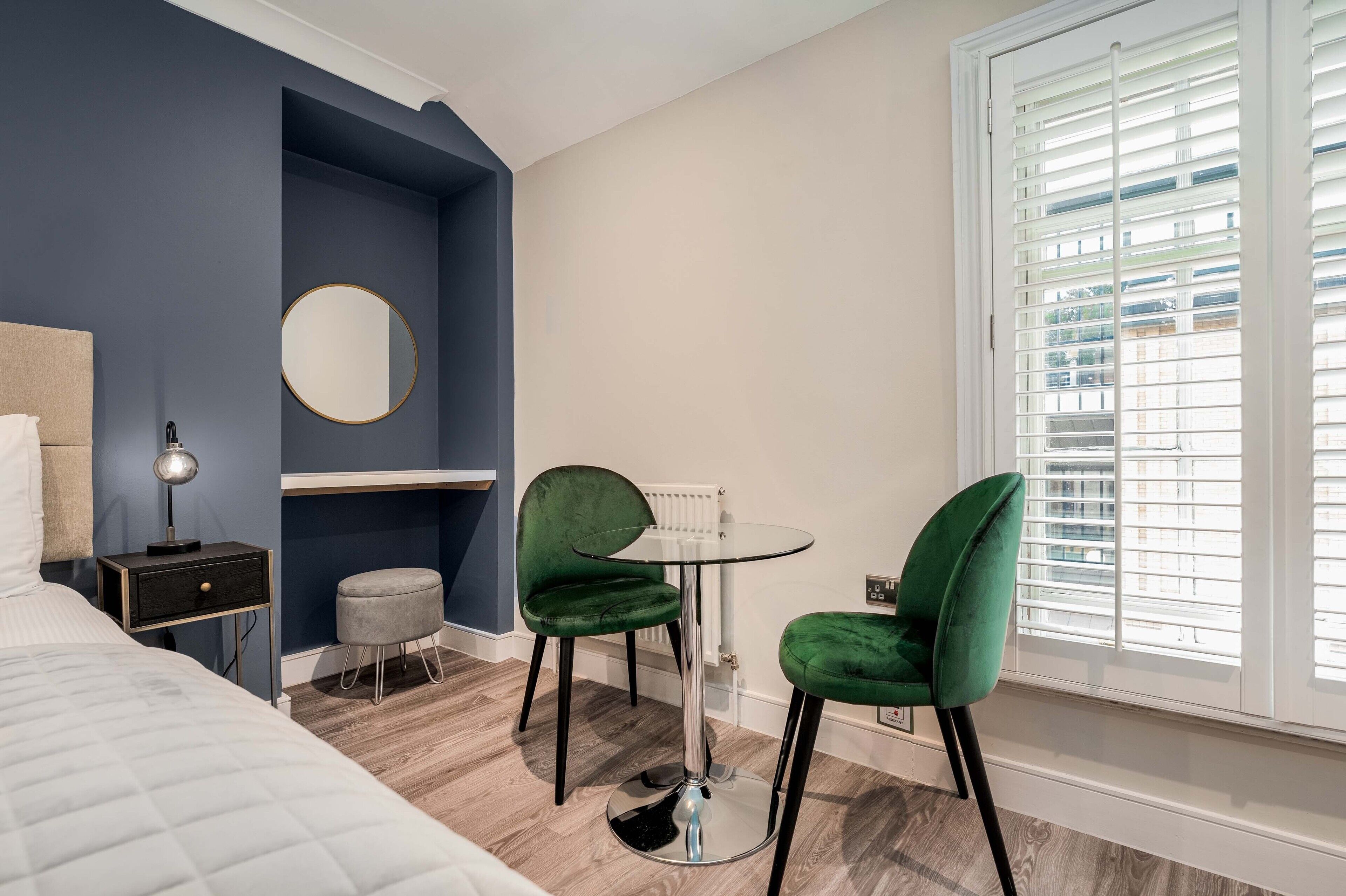 A bedroom with a bed, a desk, two green chairs, a round mirror, and a window with blinds.