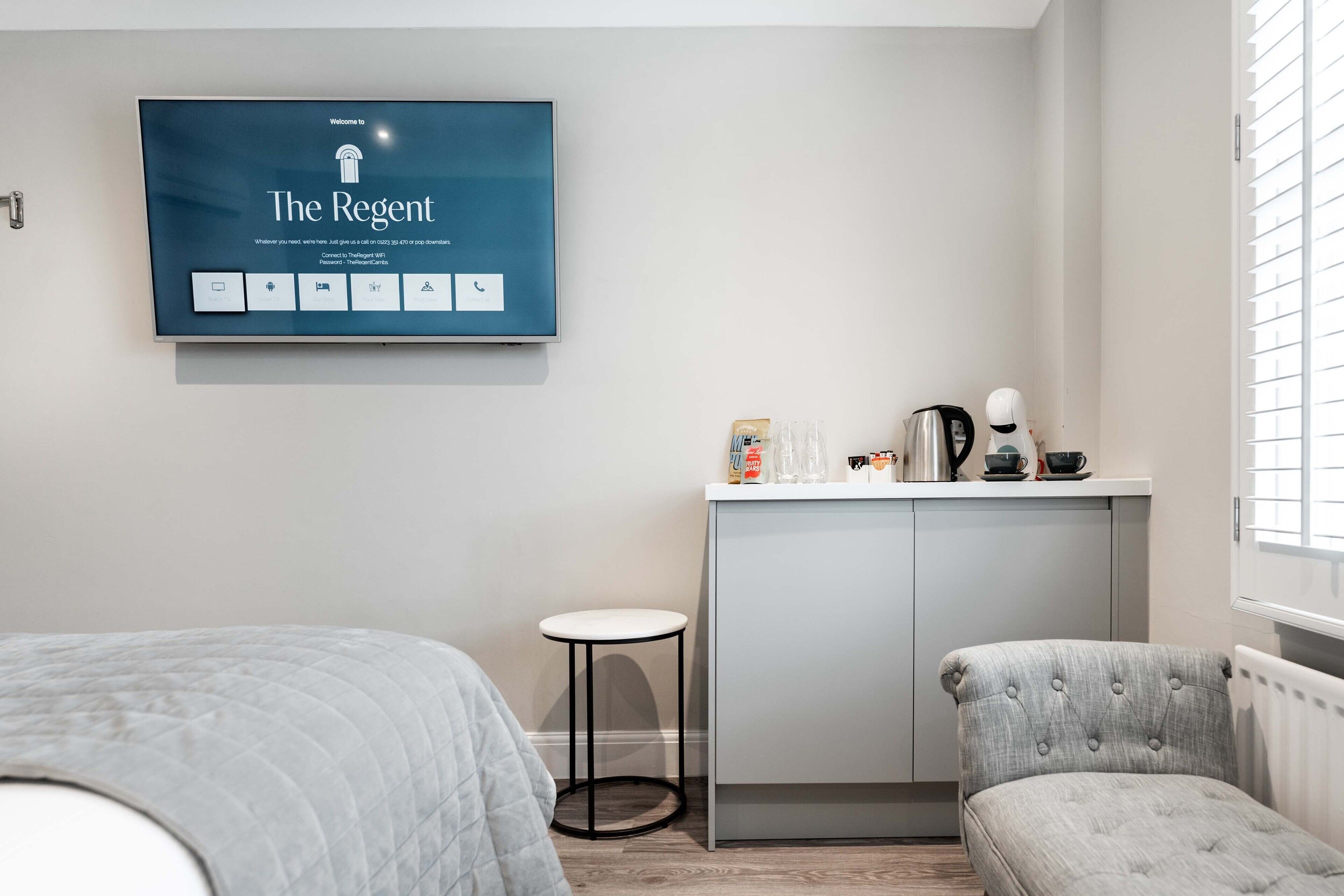 Una habitación de hotel moderna con una televisión de pantalla plana mostrando la pantalla de bienvenida 'The Regent', una kitchenette con cafetera y un área de descanso con una silla acolchada.