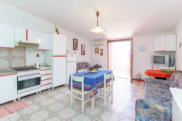 A compact kitchen with white cabinets, a gas stove, and a refrigerator. There is a dining table with a blue tablecloth and chairs. A sofa and a television are visible in the living area.