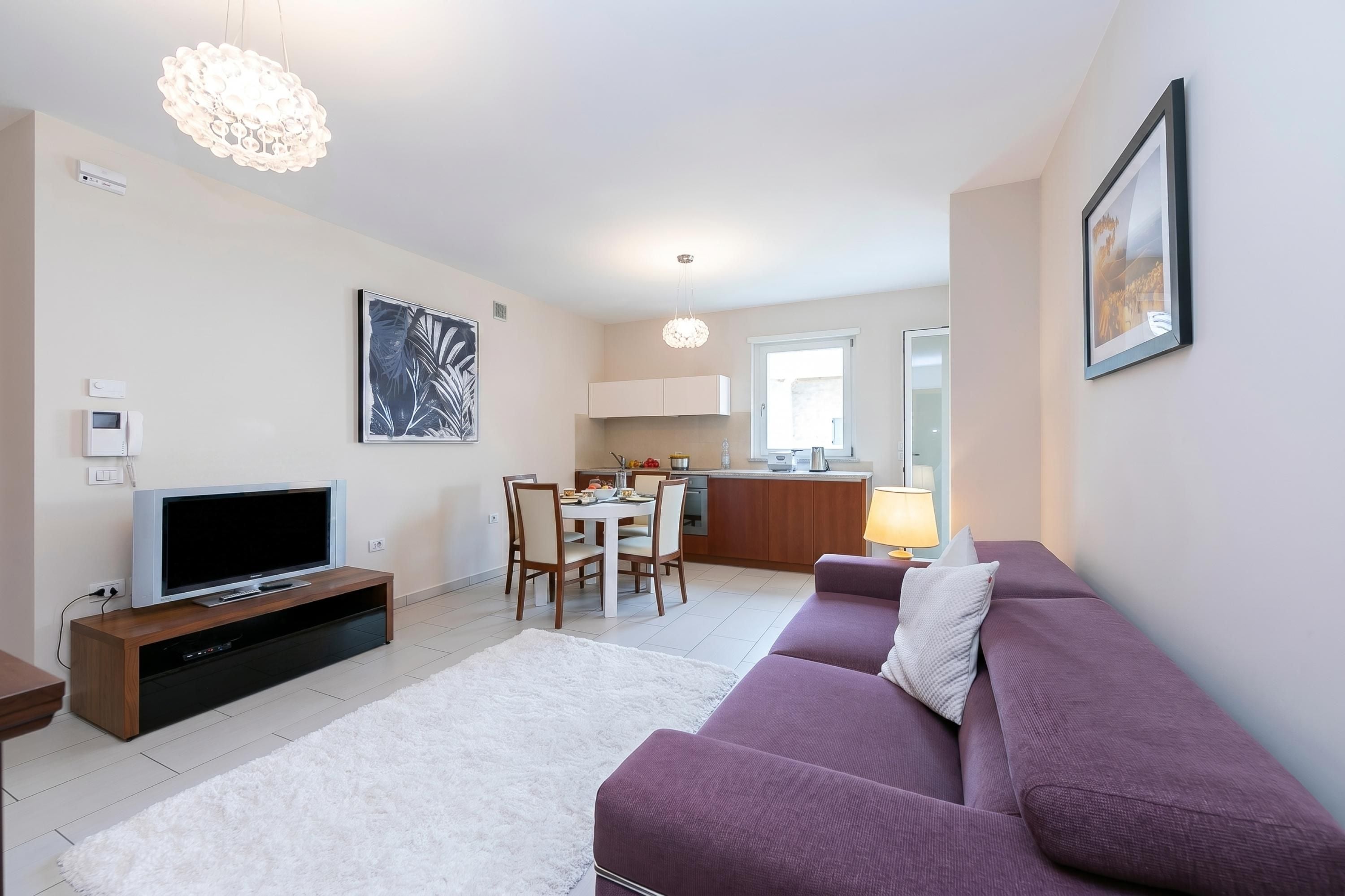 A modern living room with a flat-screen TV, a purple sofa, a dining area with chairs, and a kitchen in the background.