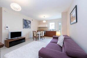 A modern living room with a flat-screen TV, a purple sofa, a dining area with chairs, and a kitchen in the background.