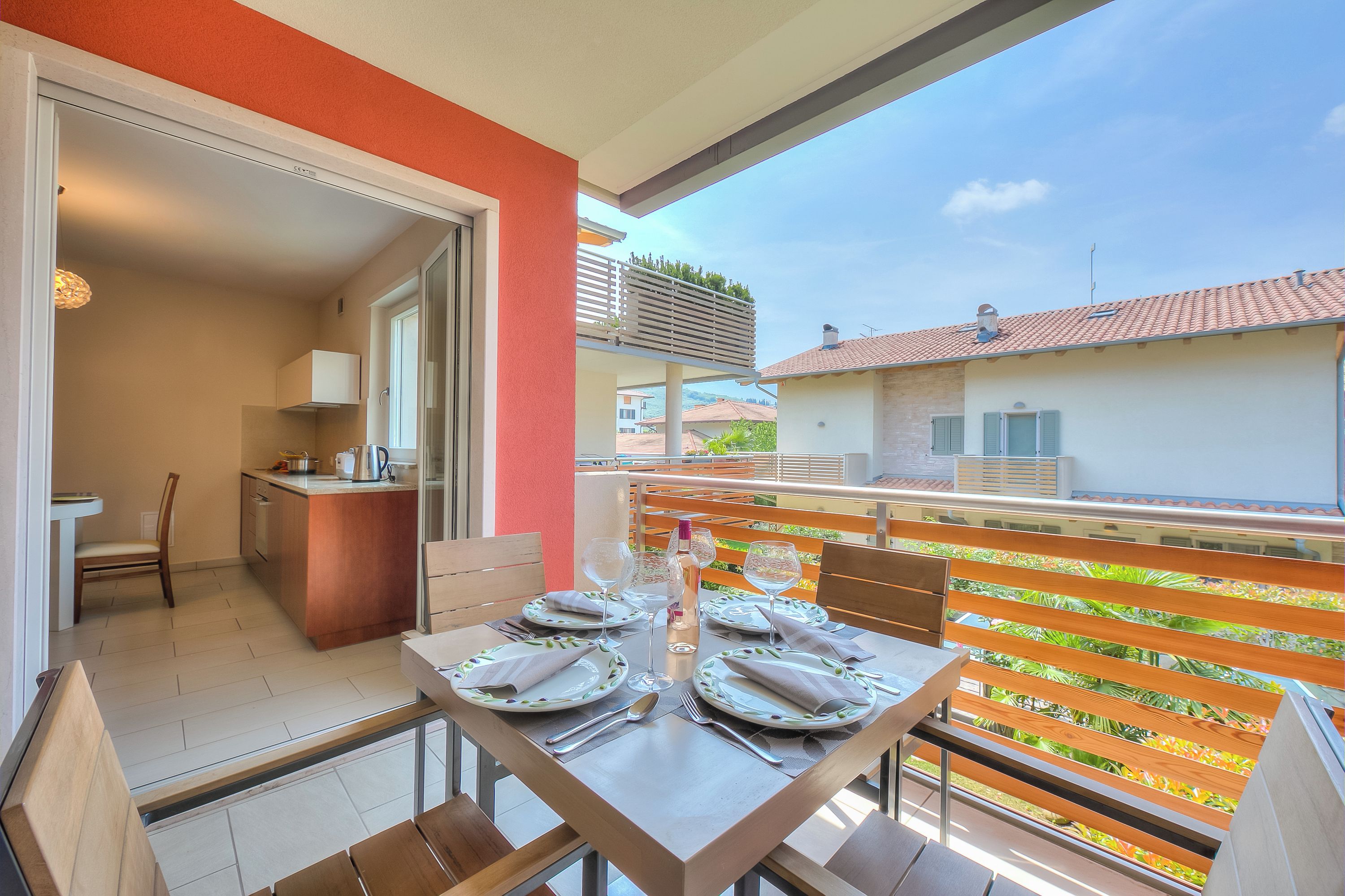 A dining area with a table set for four, a kitchen in the background, and a balcony with a view of the neighborhood.