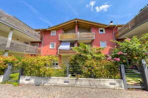 Un edificio de varios pisos con balcones, una entrada protegida por un muro y abundante vegetación.