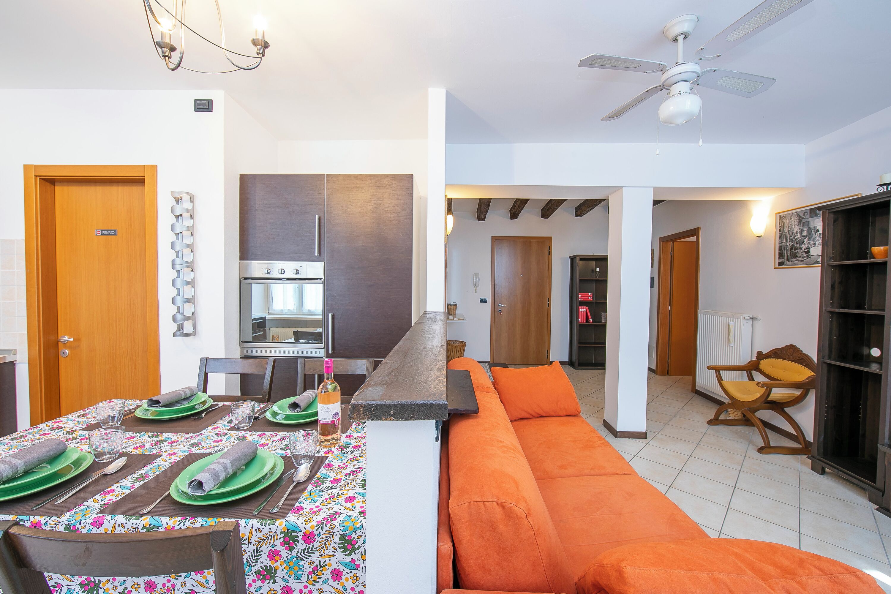 A modern kitchen with a built-in oven, a dining area with a table set for four, and a living area with a sofa and a chair.
