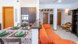 A modern kitchen with a built-in oven, a dining area with a table set for four, and a living area with a sofa and a chair. - Casa Azzurra 150m From Lake Garda (Riva del Garda)