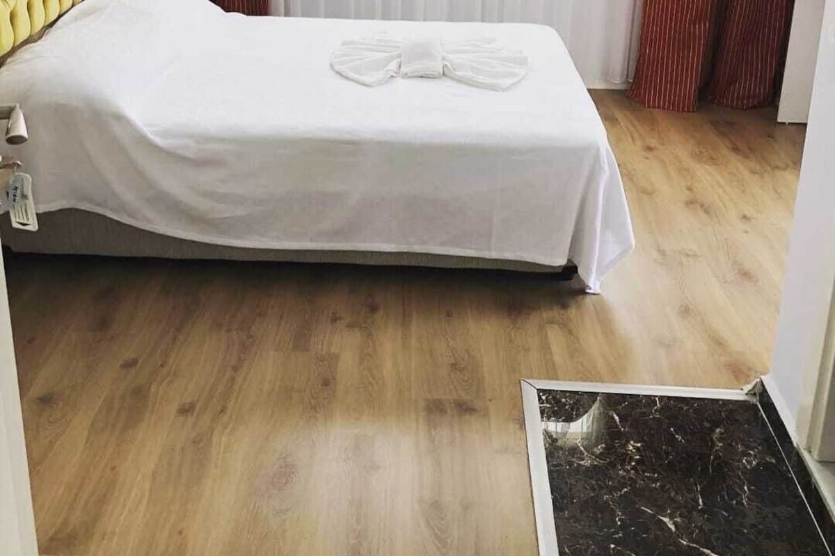 A single bed with a white bedspread and a bow tie pillow, a wooden headboard, a bedside lamp, and a view of a window with sheer curtains.