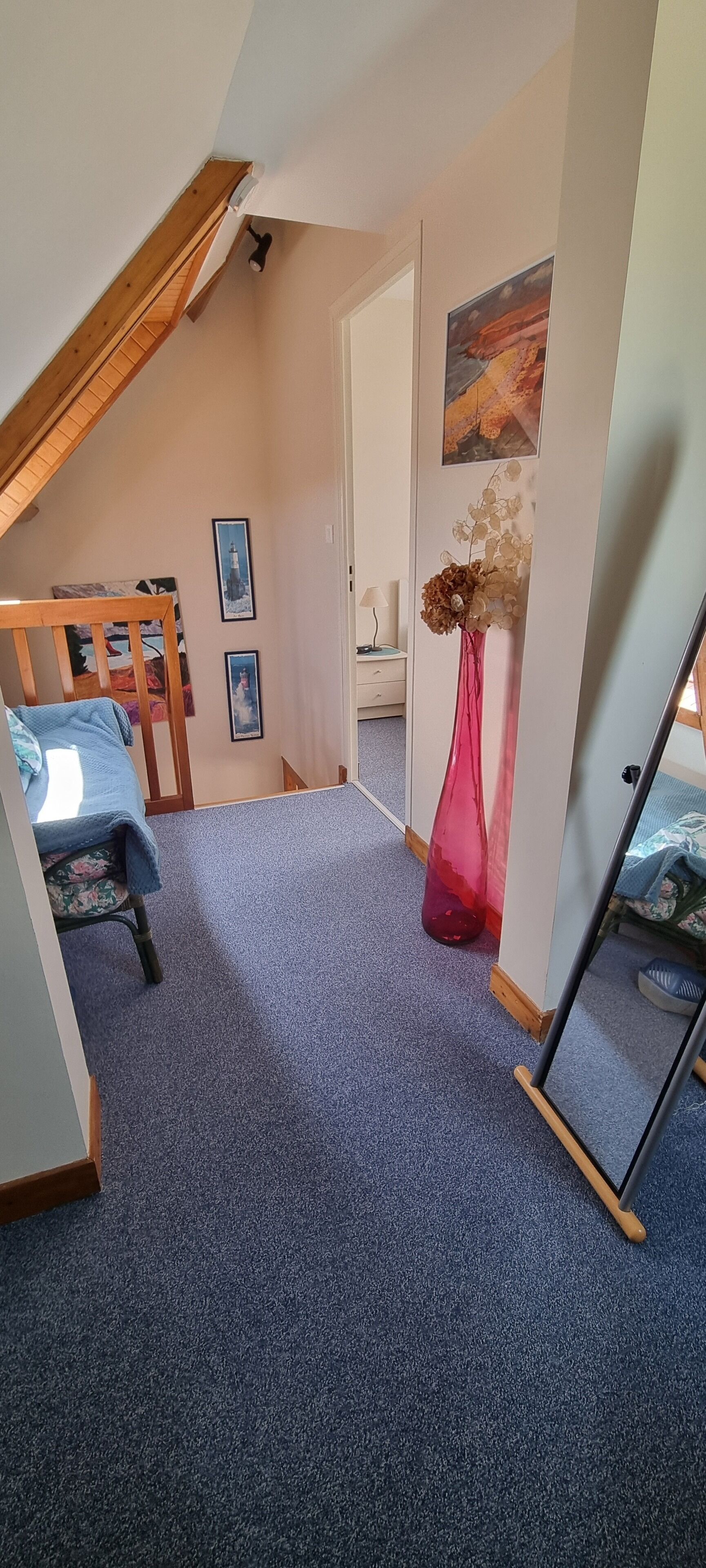 A bedroom with a sloped ceiling, a bed with a blue blanket, a red vase, and a painting on the wall.