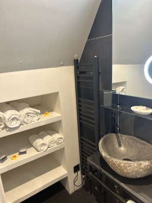 Un baño moderno con un lavamanos de piedra grande, estantes blancos con toallas y un toallero negro.