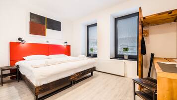 A modern bedroom with a red headboard, a wooden bed frame, a small bedside table, a desk with a chair, and two windows with blinds.