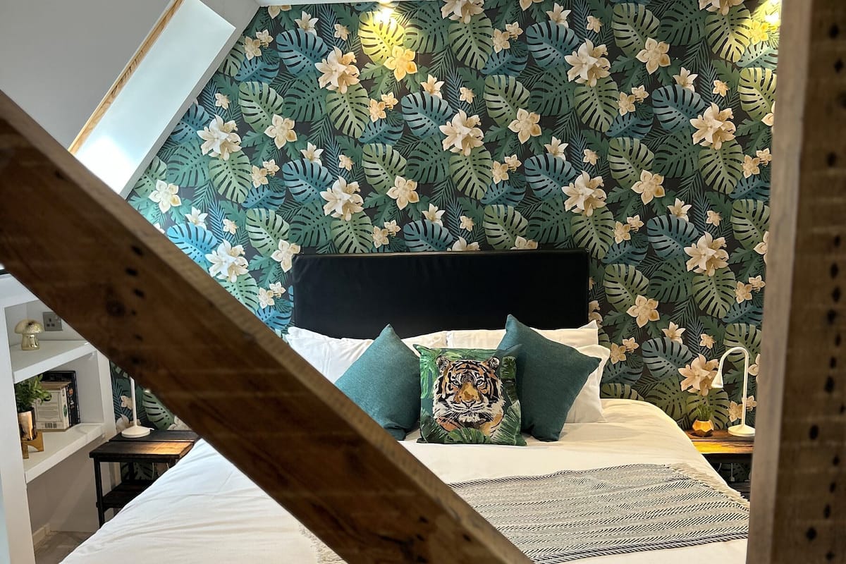 A bedroom with a bed, pillows, and a headboard against a wall with a tropical leaf and flower wallpaper.