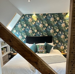 Un dormitorio con cama, almohadas y cabecera frente a una pared con estampado de hojas y flores tropicales.