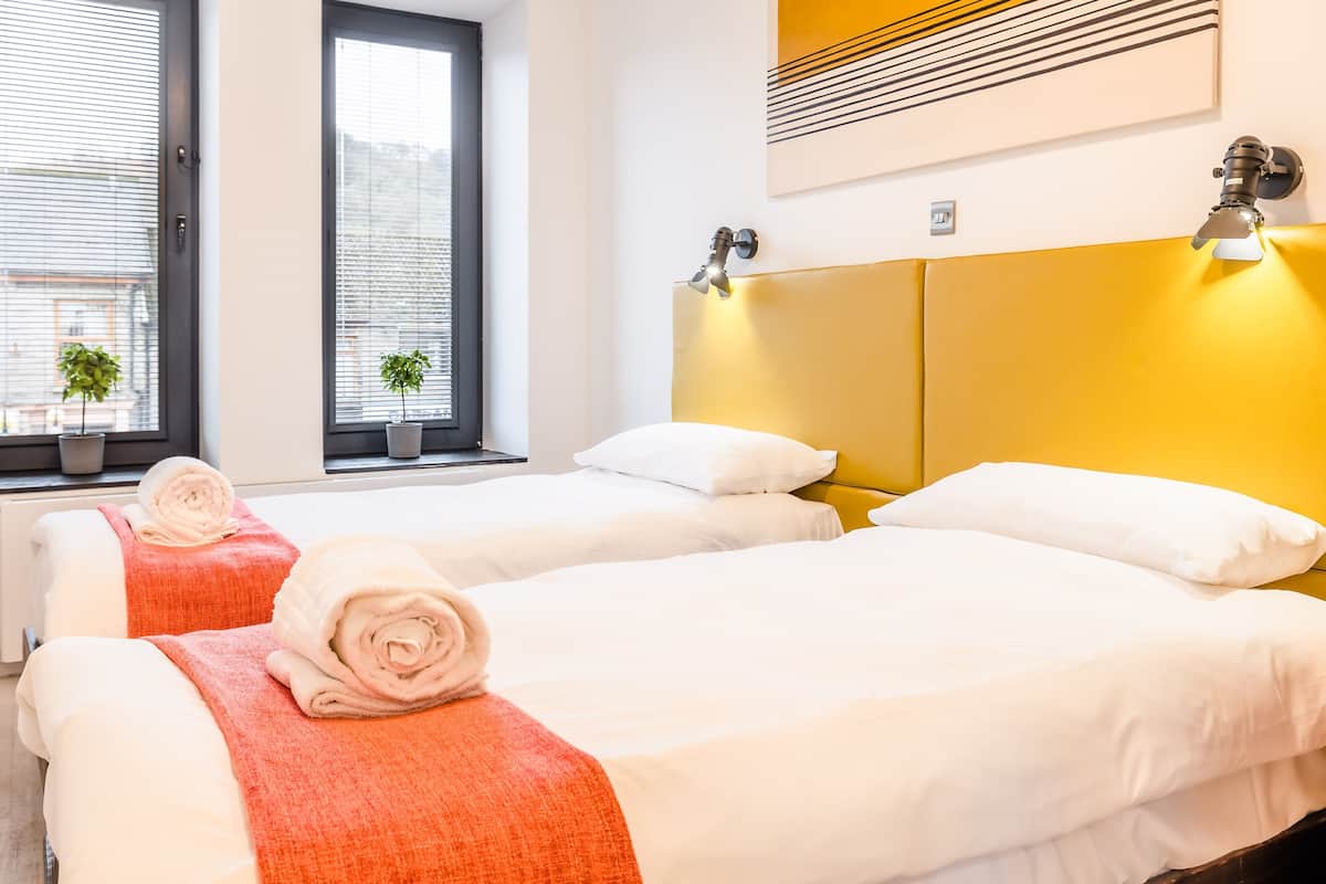 A hotel room with two beds, a yellow headboard, and a window with blinds.