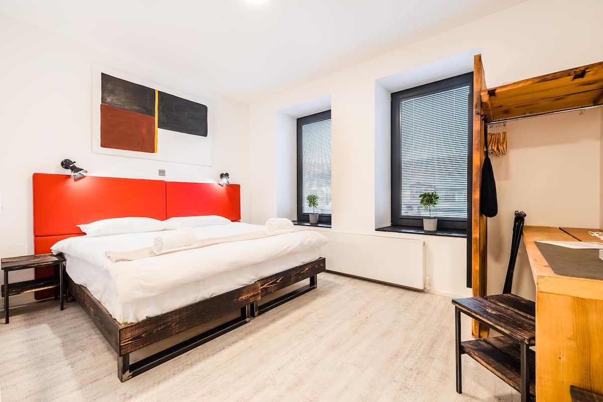 A modern bedroom with a red headboard, a wooden bed frame, a desk, and two windows with blinds.