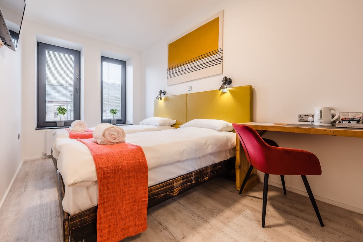 A bedroom with a bed, two windows, a desk, a chair, and a painting on the wall.