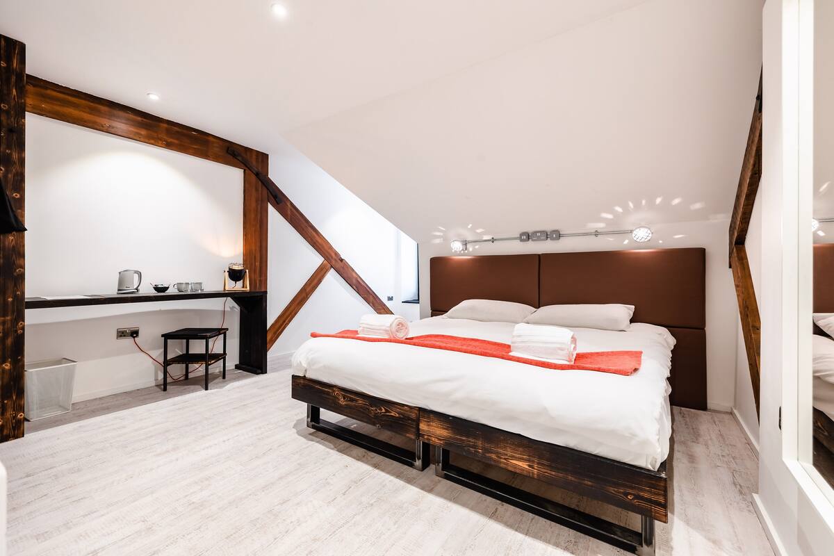 A modern bedroom with a large bed, wooden beams, a desk, and a small stool.