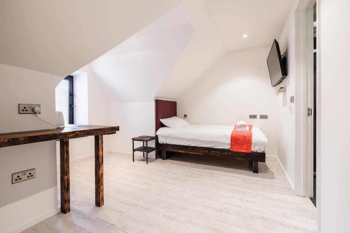 A bedroom with a sloped ceiling, a bed with a white mattress and a red blanket, a wooden nightstand, and a TV mounted on the wall.