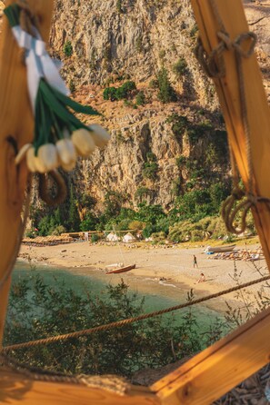 A view of a beach with a rocky cliff in the background, a wooden structure in the foreground, and a small boat on the water. - Butterfly Valley Seaside Glamping (Fethiye)