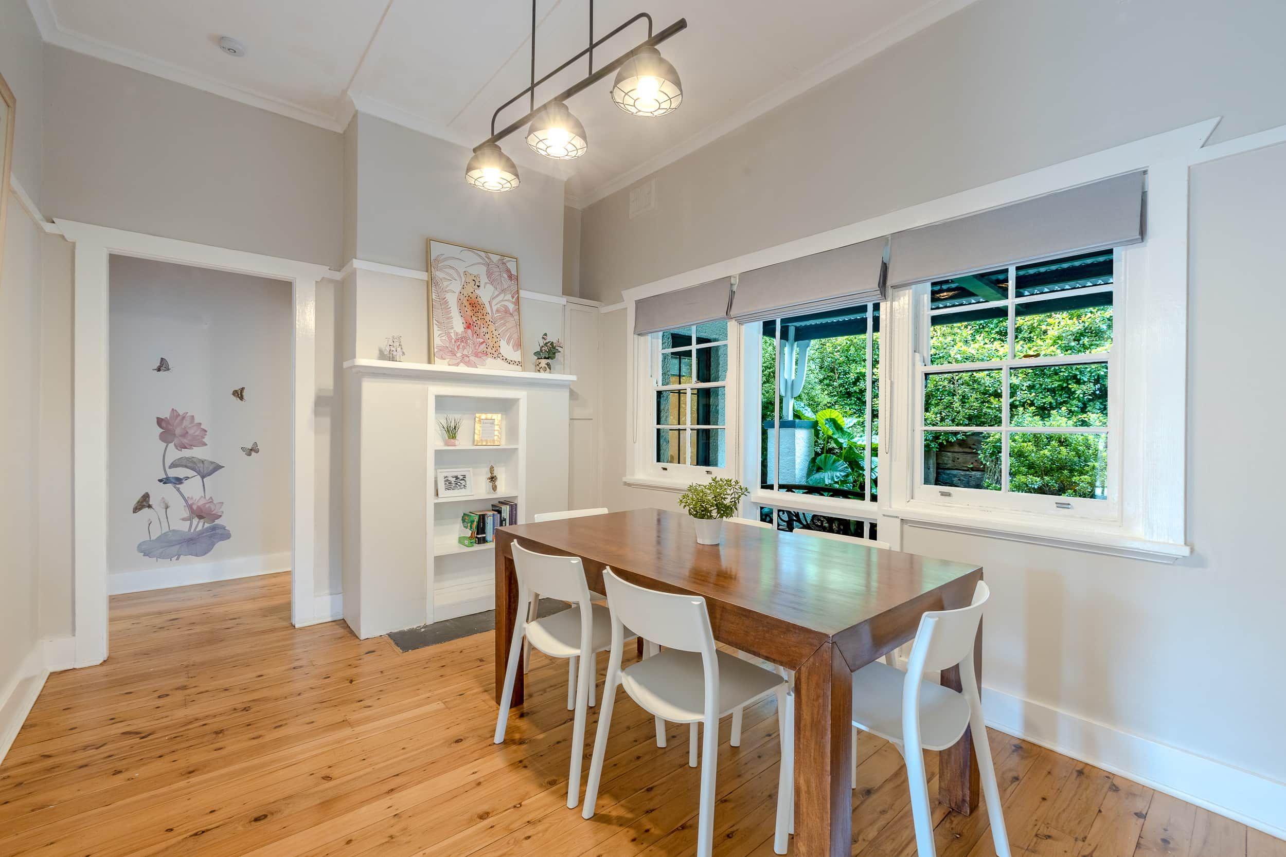 A dining area with a wooden table and white chairs, a white fireplace with a mantel, and a large window with a view of greenery.