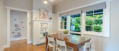 A dining area with a wooden table and white chairs, a white fireplace with a mantel, and a large window with a view of greenery.