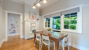 A dining area with a wooden table and white chairs, a ceiling light fixture, and a window with a view of greenery. - Sunshine Cottage with private garden, Katoomba (Katoomba)