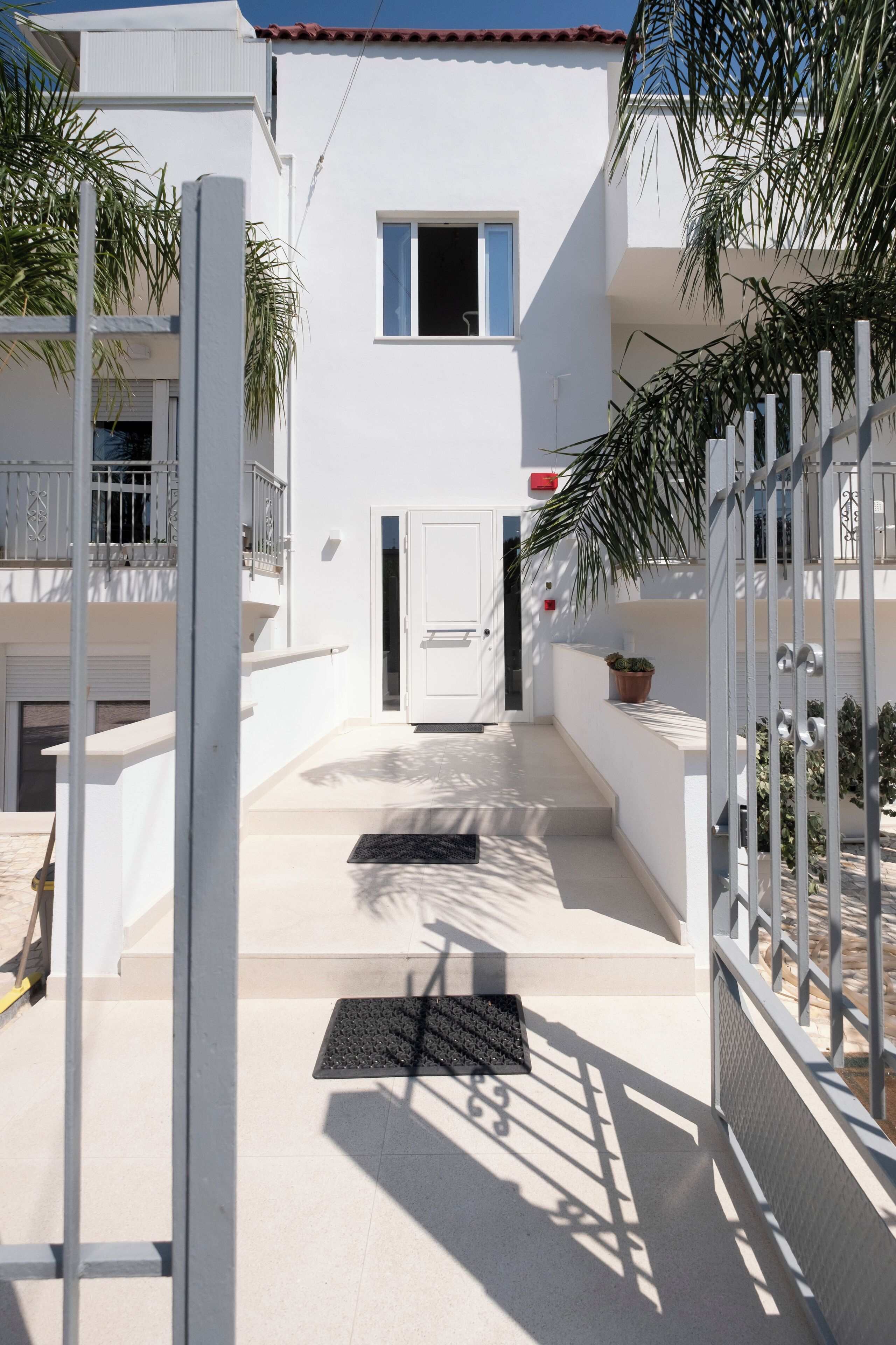 Un edificio blanco con techo de tejas rojas, una puerta y un ventanal pequeño. Hay escalones que conducen a la puerta y una verja metálica a la derecha.