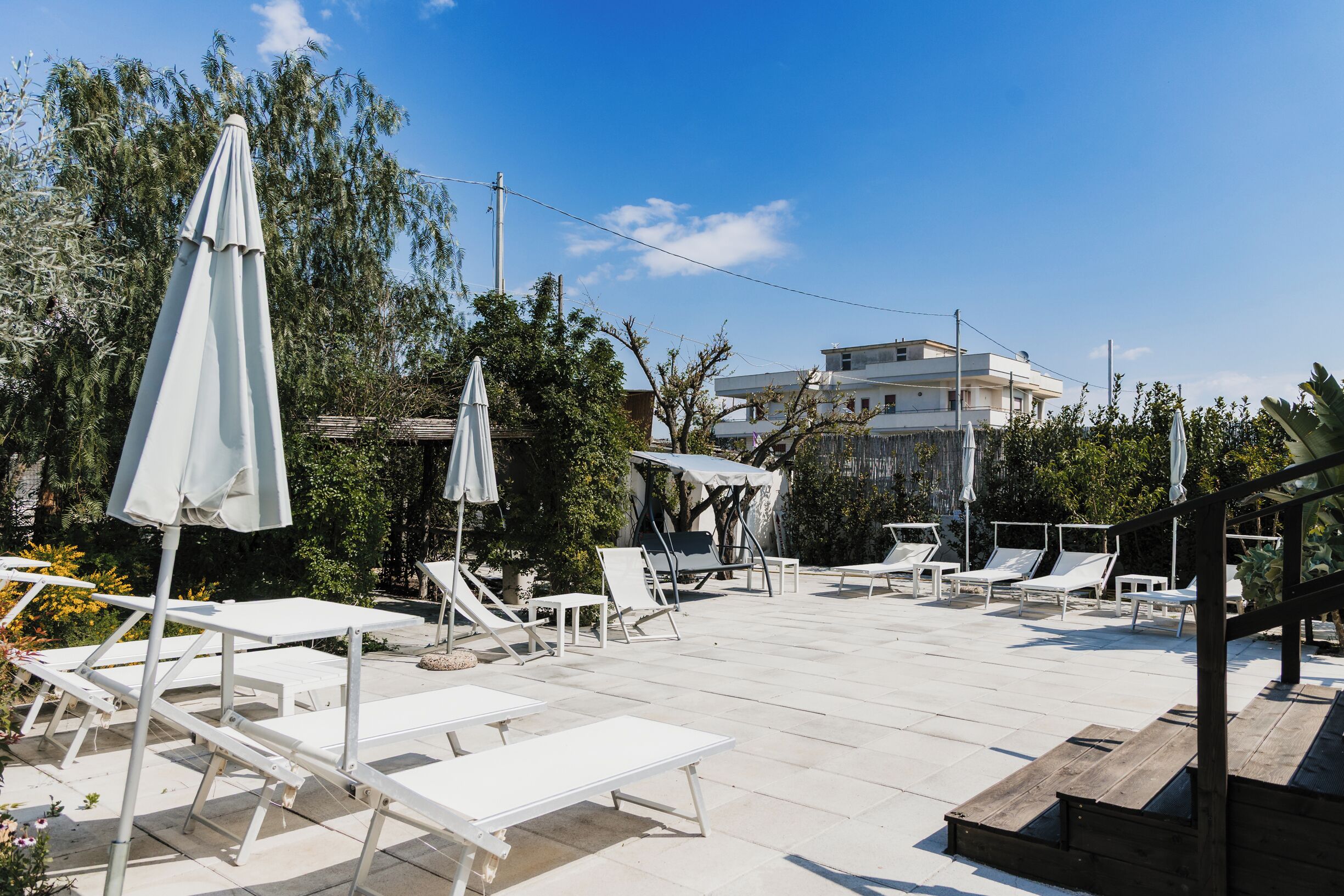 A rooftop pool area with white lounge chairs and umbrellas, surrounded by trees and a building in the background.