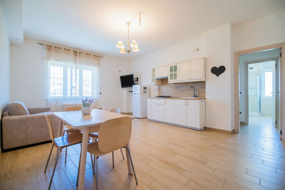 A modern kitchen with white cabinets, a dining area with a table and chairs, and a living area with a sofa.