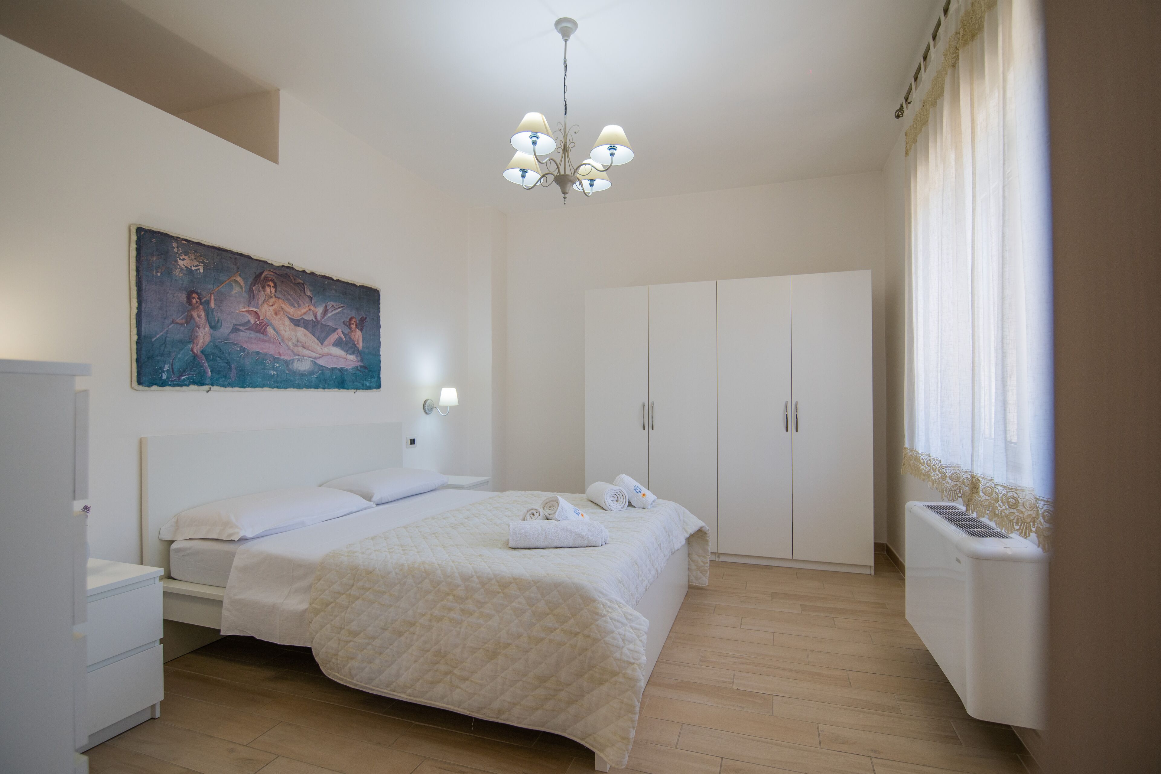 A bedroom with a bed, a nightstand, a wardrobe, a painting on the wall, and a ceiling light.