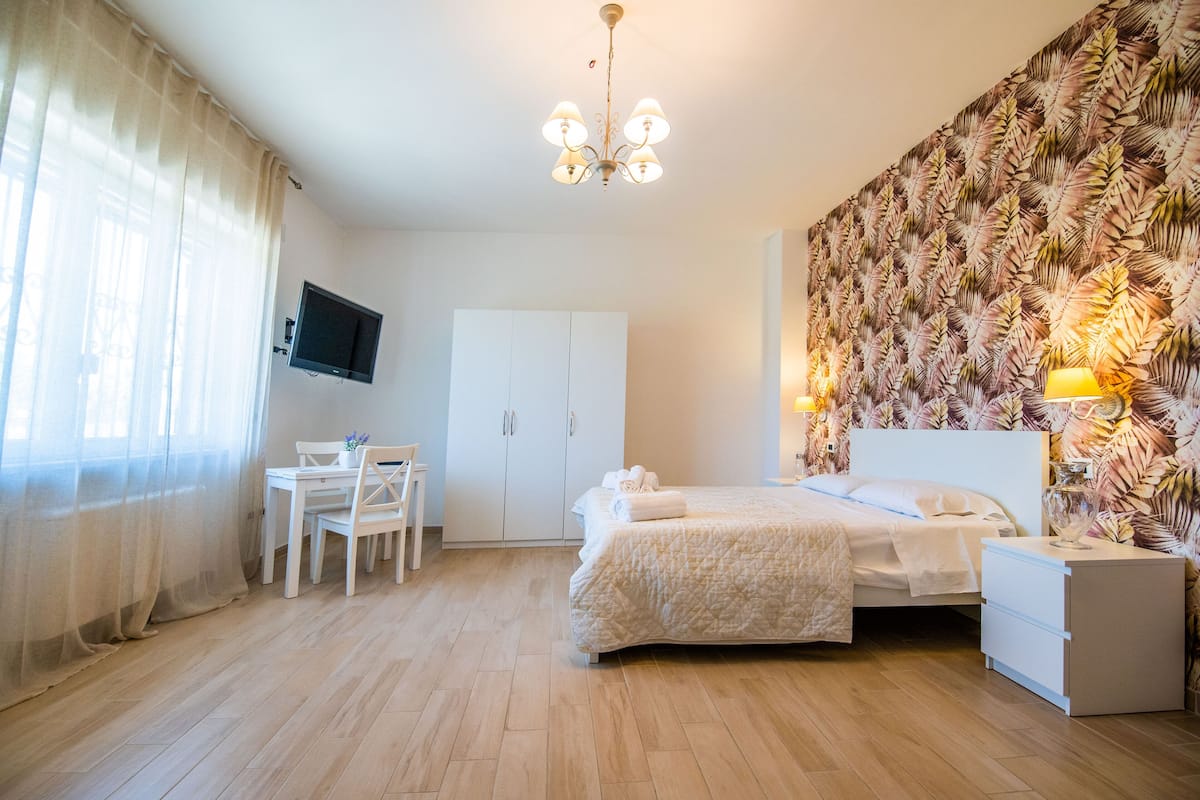 Ein Schlafzimmer mit Bett, Nachttisch, Schreibtisch, Stuhl, Fernseher, Kleiderschrank und einer Wand mit Blättermuster.