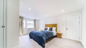A bedroom with a large bed, a wooden nightstand, a white door, and a window with curtains. - Livestay - 2 Bed Hollywood Movie Themed Apt (Barnet)