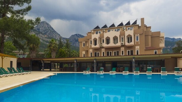 A swimming pool with lounge chairs, a building with multiple balconies, and mountains in the background. - Alexius Beach Hotel (Kemer)