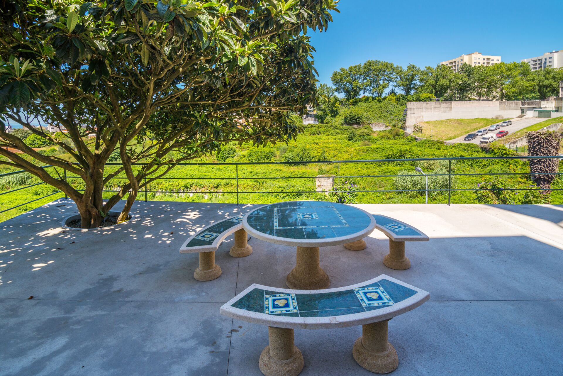 A round table with a tiled top, surrounded by a low stone wall, set on a concrete surface with a tree in the background.