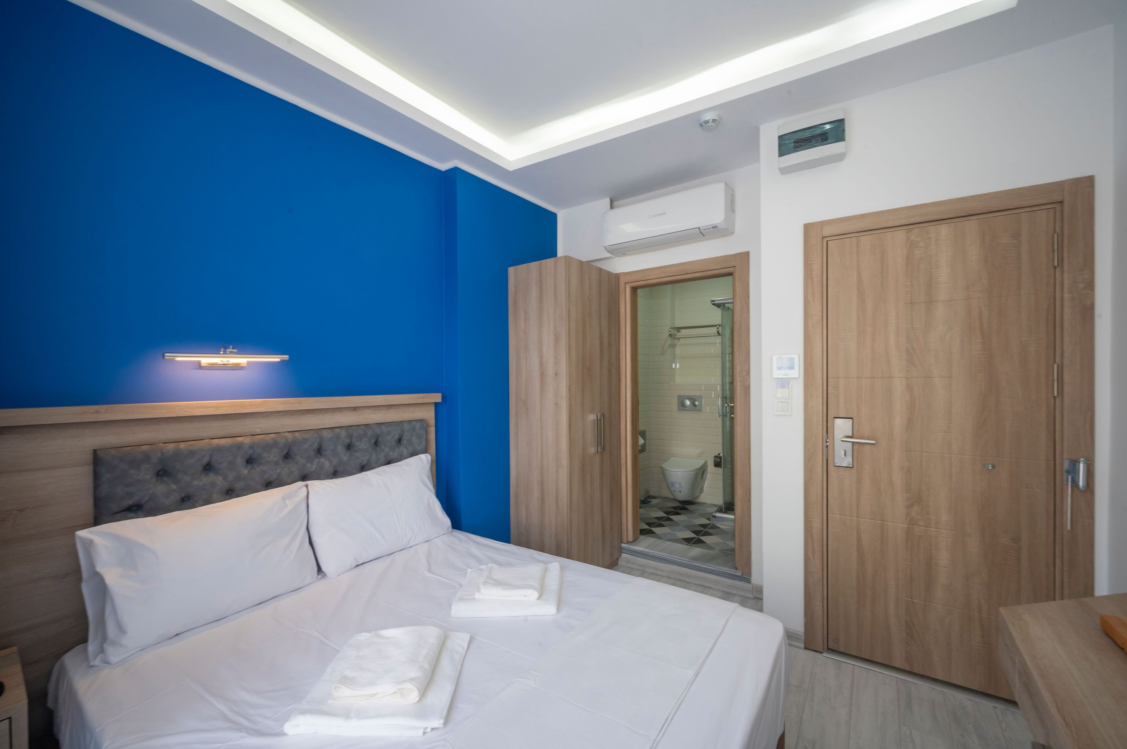 A hotel room with a bed, a wooden wardrobe, a bathroom with a shower, and a blue accent wall.