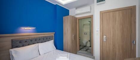 A hotel room with a bed, a wooden wardrobe, a bathroom with a shower, and a blue accent wall.